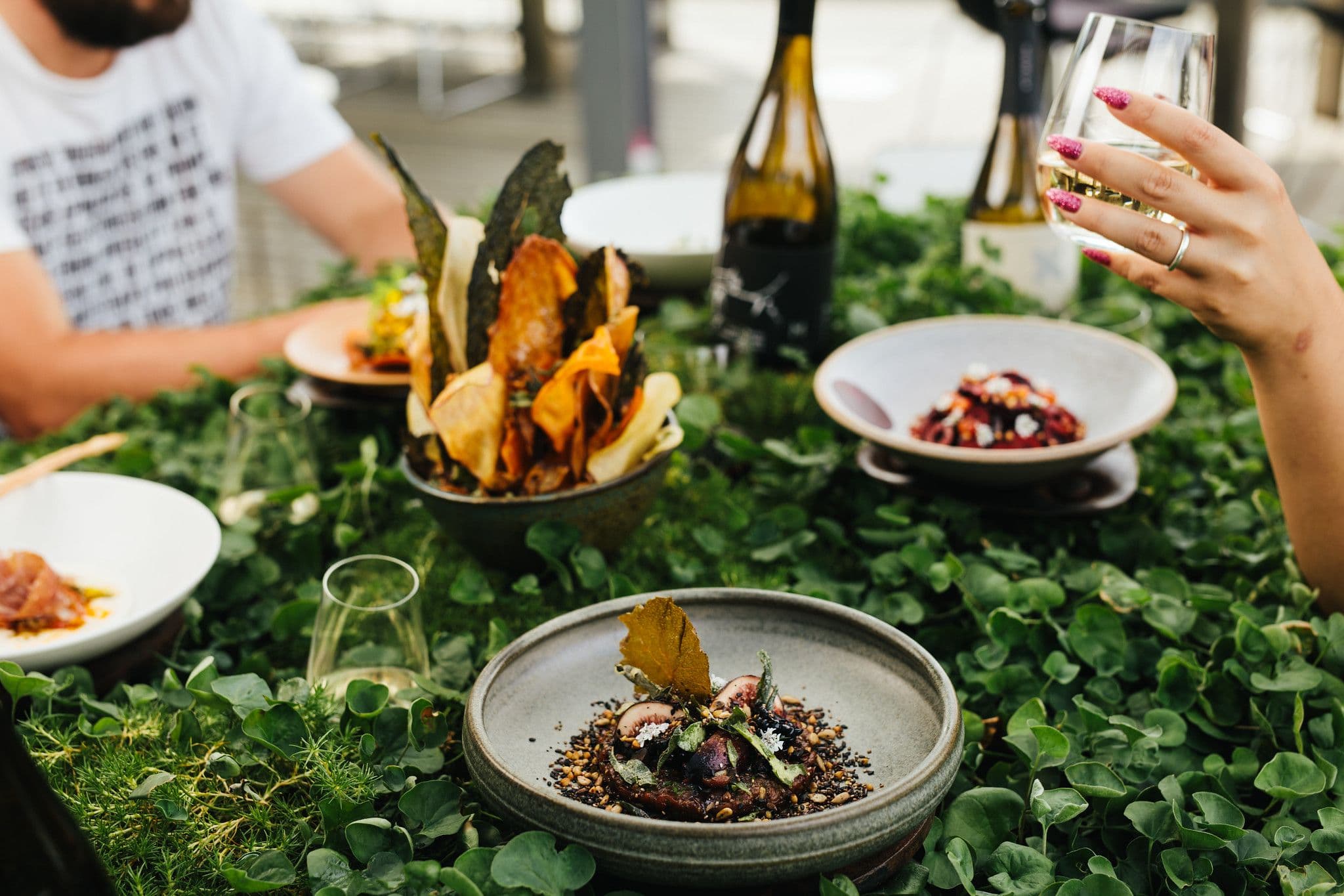 People dining on a lush tabletop of leaves with ceramic plates containing vibrant thoughtfully plated food.