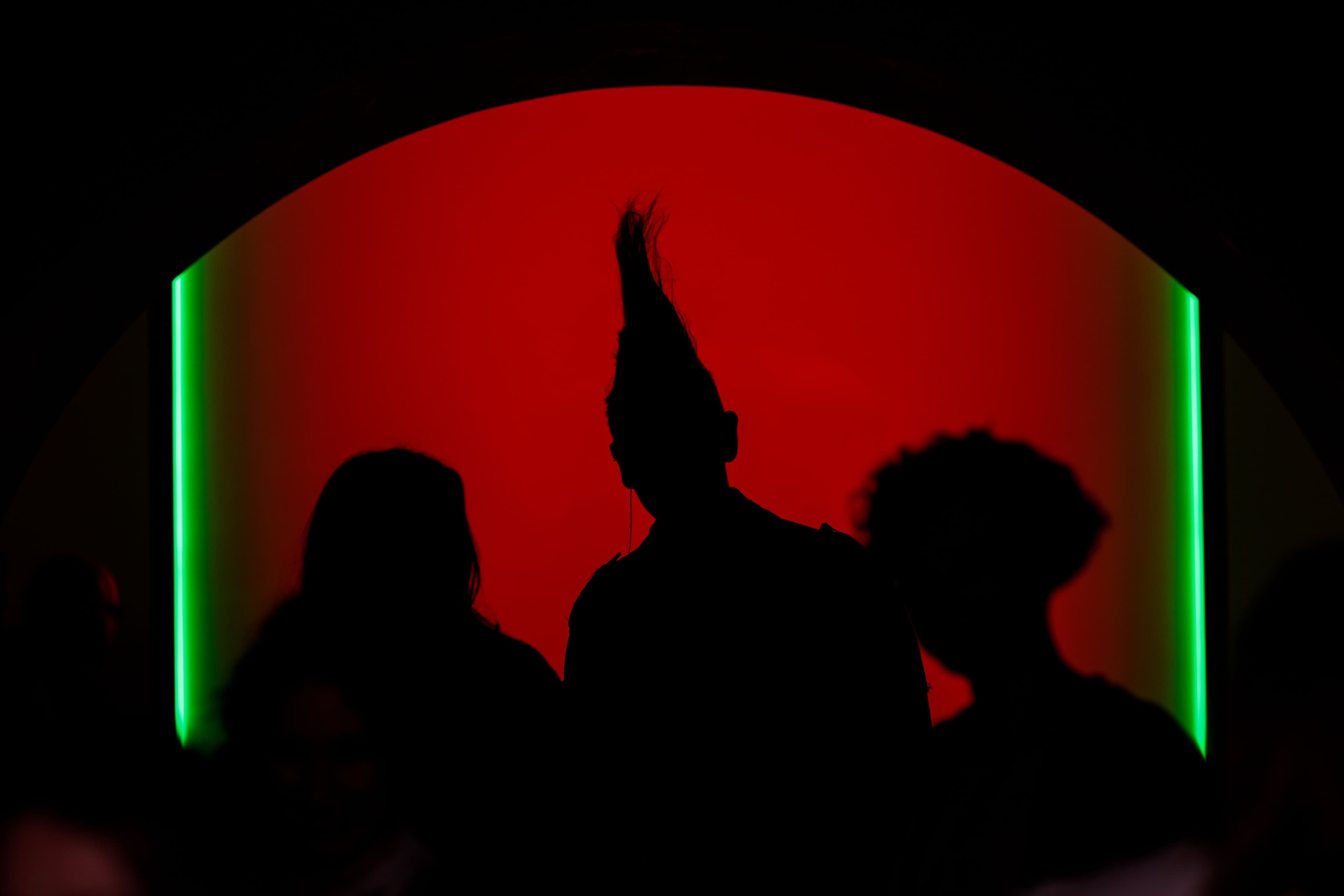 Silhouttes of three figures, one with a mohawk standing in front of a background of red light with green vertical strips at the sides