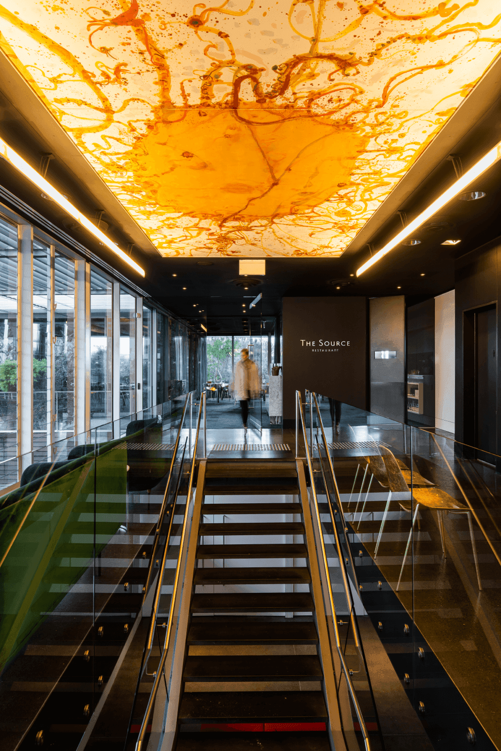 The entrance of the Source restaurant, featuring a staircase and a large abstract painting of the sun, illuminated from above.