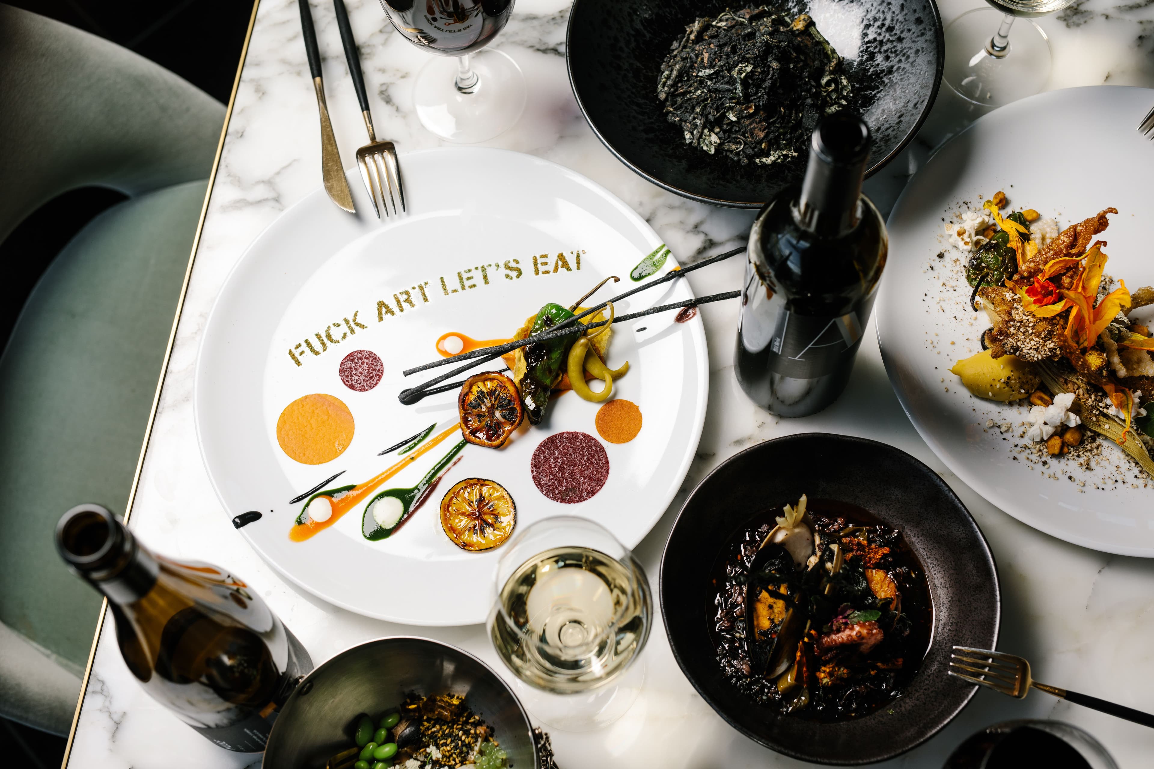 A table scene with "FUCK ART LET'S EAT" stenciled onto a plate