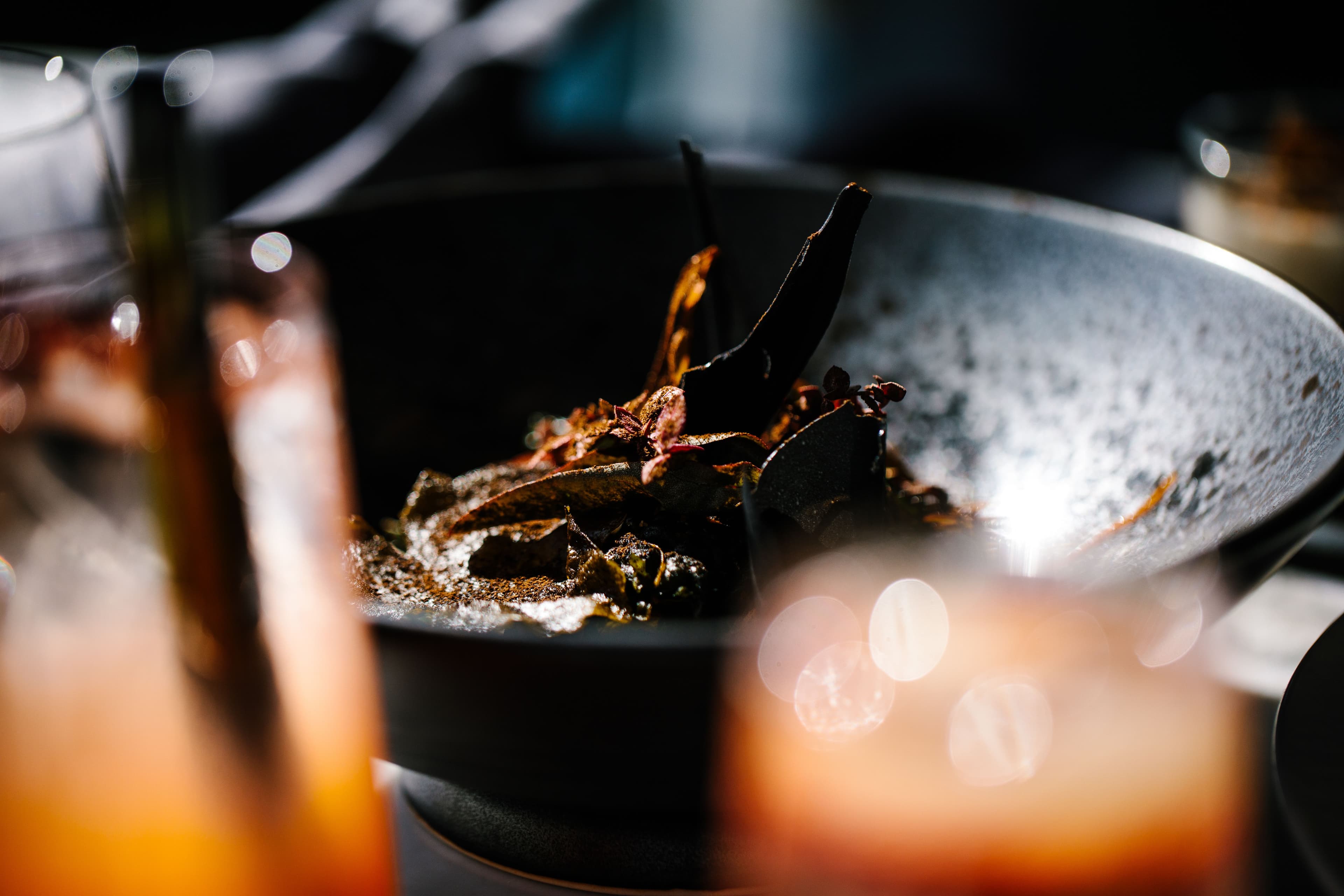 A dimly lit image of a bowl of structurally garnished food with orange cocktails in soft focus in the foreground