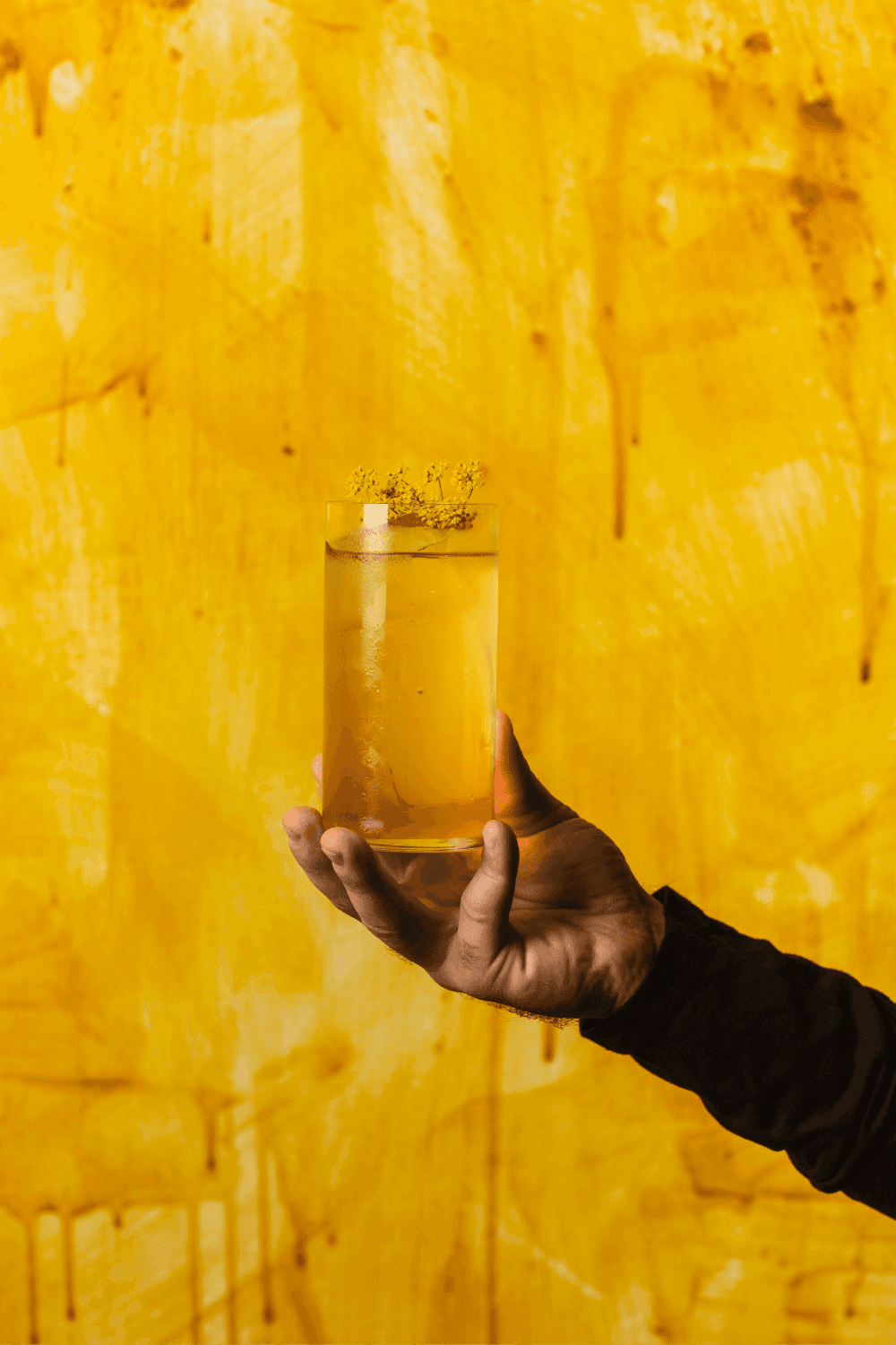 A hand holding a high ball cocktail in front of a yellow wall