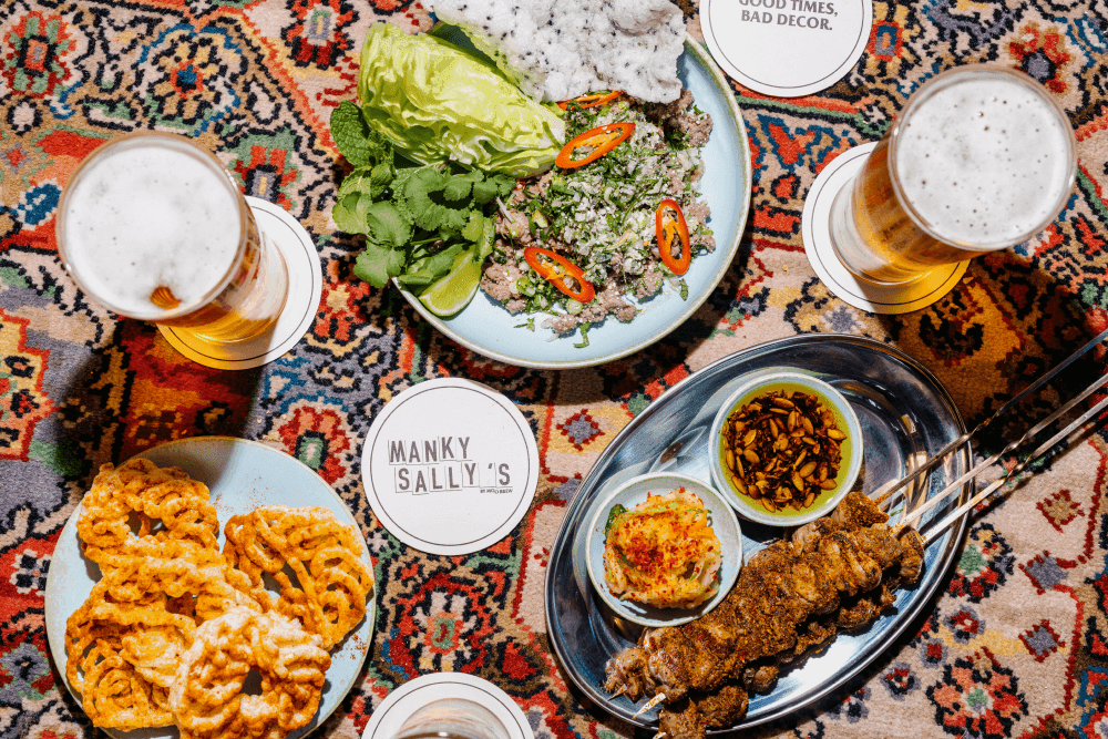 Image of a vibrant patterned carpet with Manky Sally's branded coasters, beers and a selection of crispy, spicy, salty looking snacks and a vibrant fresh salad.