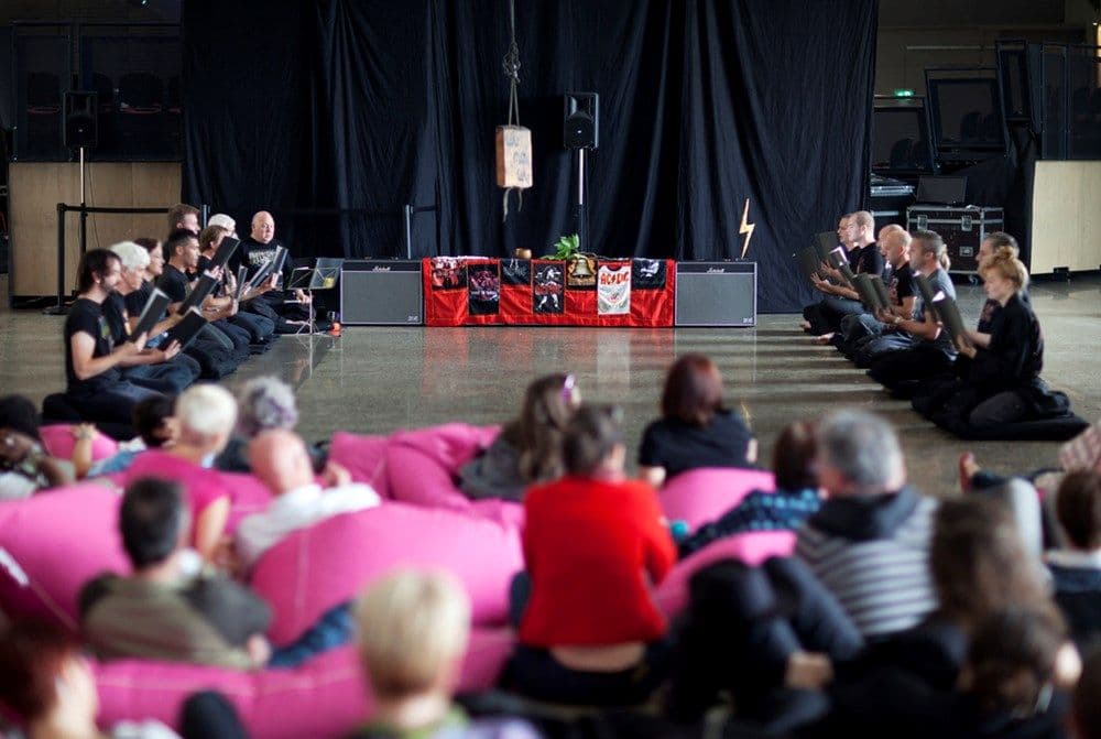 Two rows of people chanting from books infront of an altar of Amplifiers. Audience watches in the foreground.